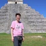 Eine hellhäutige Frau mit dunklen, kurzen Haaren, einer Brille, einem rosafarbenen T'Shirt und dunkelgrauen Hosen steht vor einer Maya-Pyramide in Chichen-Itza